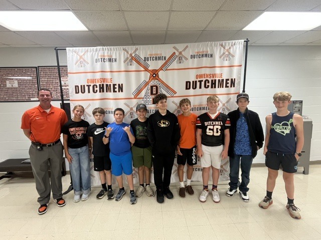 Nine young boys and a man stand in front of a Dutchmen banner in a room.