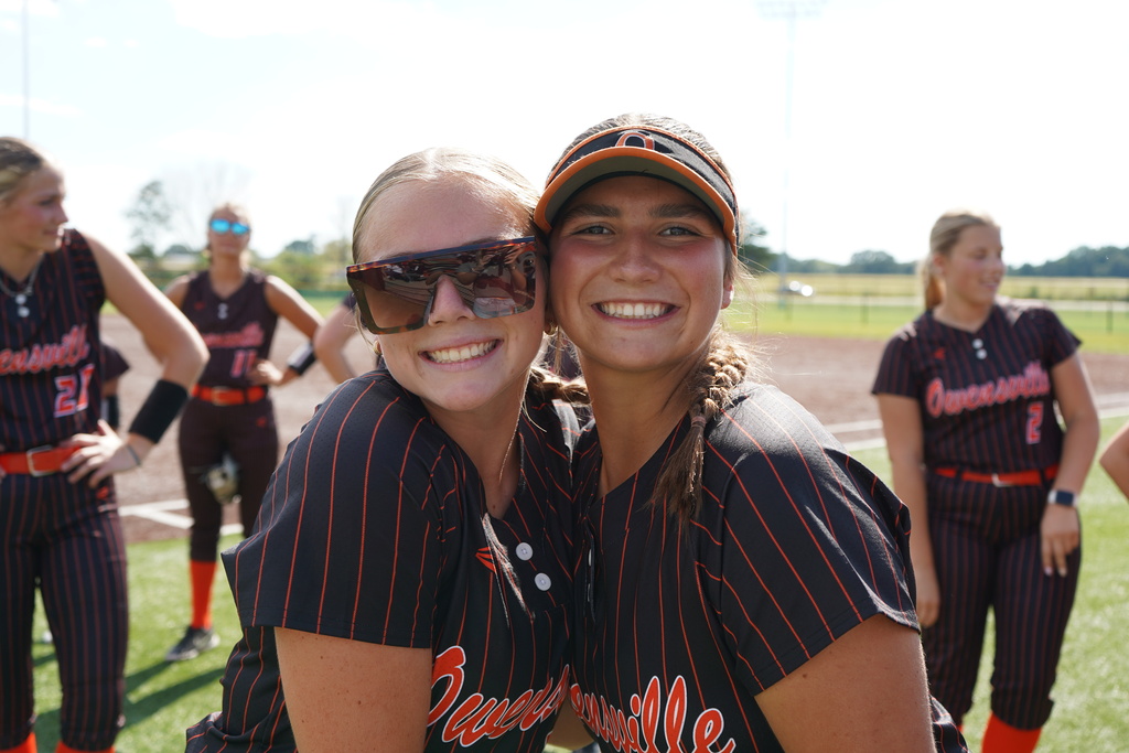 Two girls in matching striped jerseys and hats pose for a photo on a baseball field.