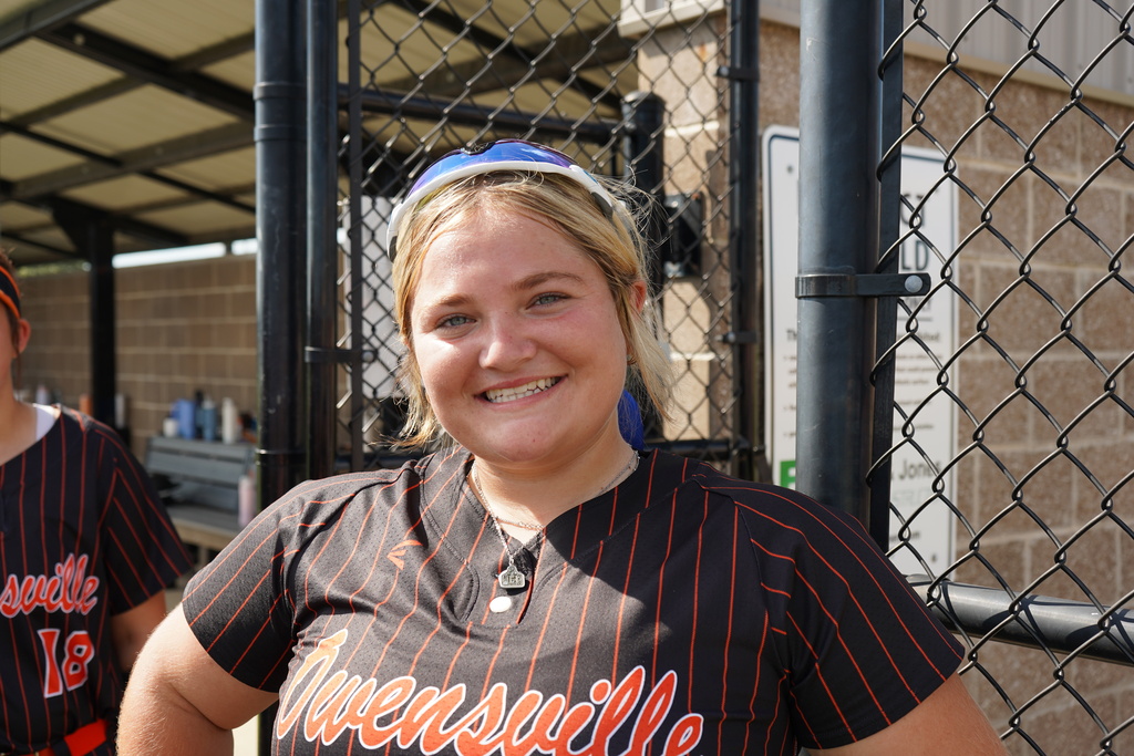 A smiling woman in a striped uniform stands near a baseball dugout with a fence.