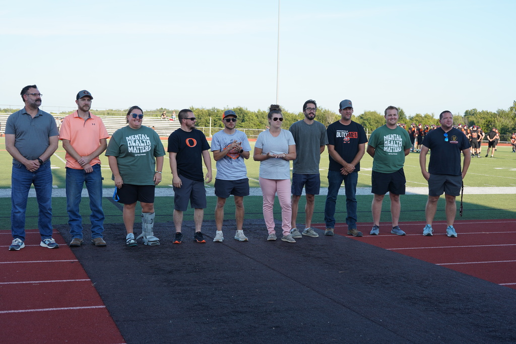 Group of people in athletic wear and caps stand on track, some wearing sunglasses, in front of an outdoor field.