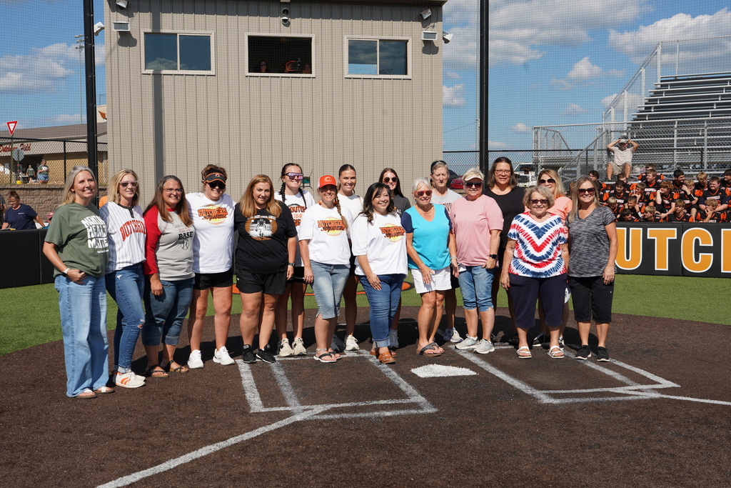Group of people wearing shirts, standing on a baseball field in front of a building.