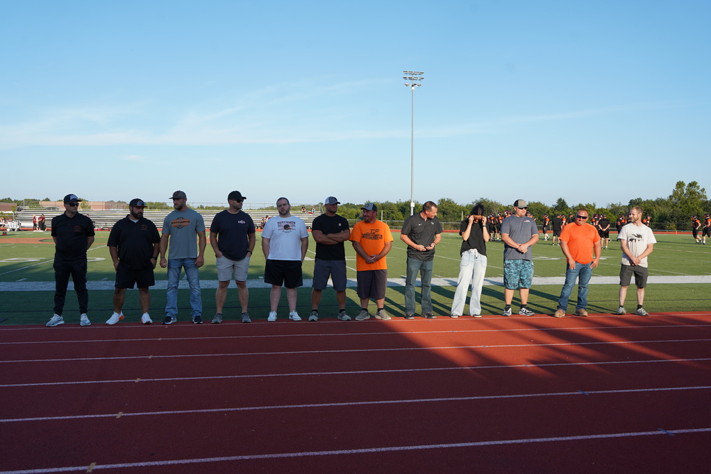 A group of people stand on a red track, some wearing hats and sneakers, with a grassy field and trees in the background.
