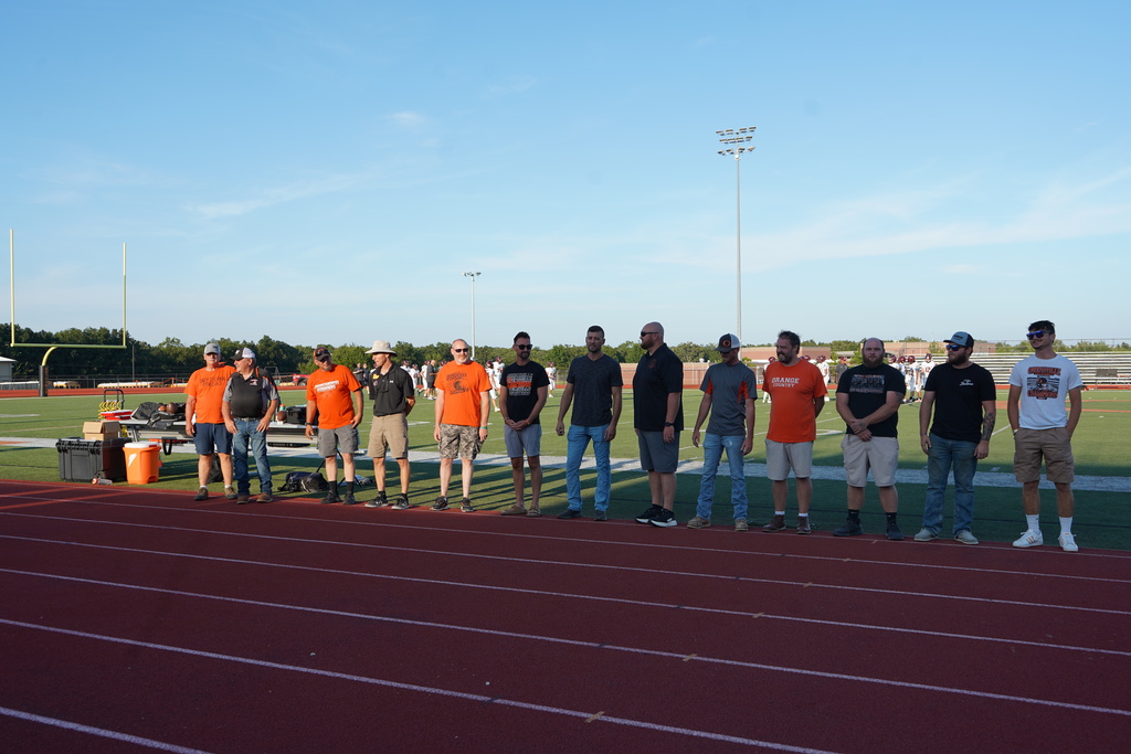 Eleven individuals stand in a row on a track. Most wear orange shirts; a few wear black. Behind them is a football field.