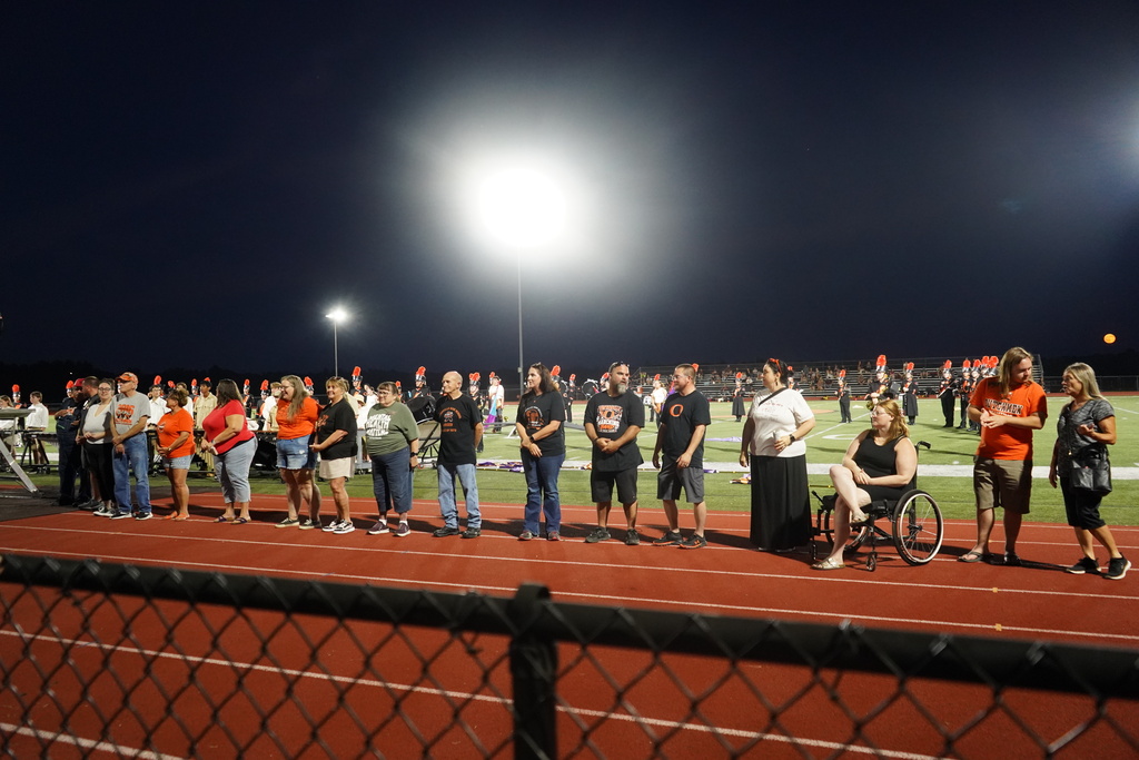 People in various attire stand in a line on a track with a fence, illuminated by streetlights.