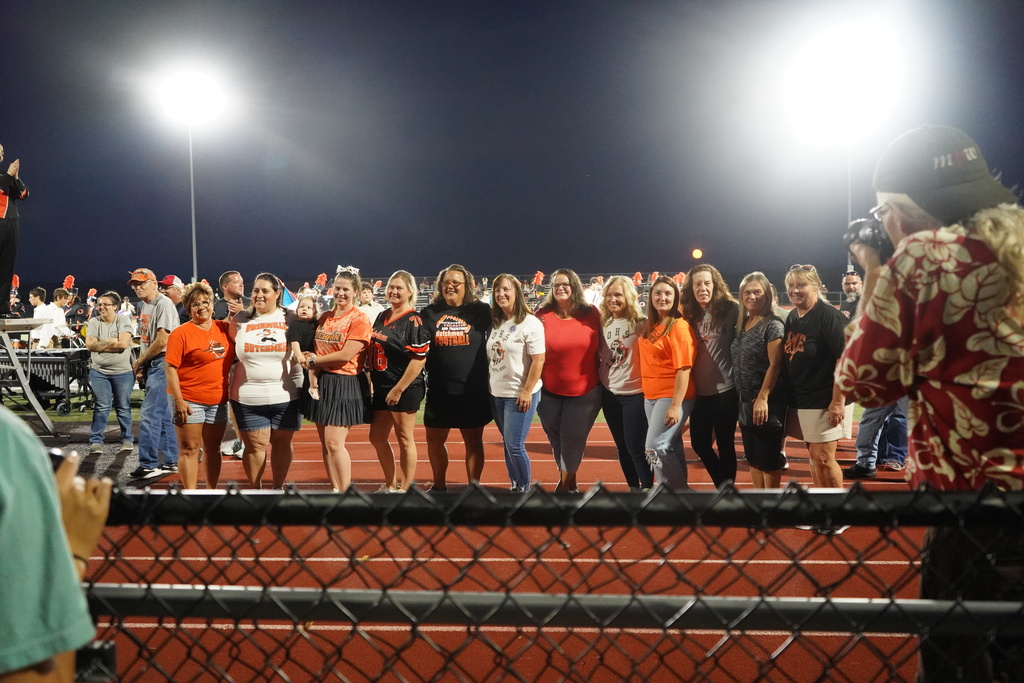 A group of people, wearing various colored shirts, stands on a track field at night. A person with a camera captures the scene.