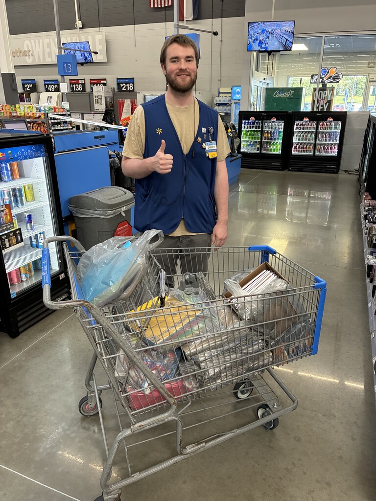 Person in blue vest gives a thumbs up in a store, standing next to a full shopping cart.