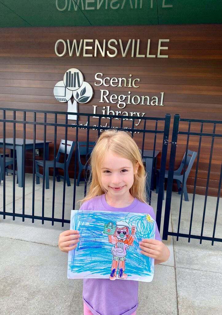 A child holds a drawing in front of a black fence. Behind the fence, a library sign reads "Scenic Regional Library."