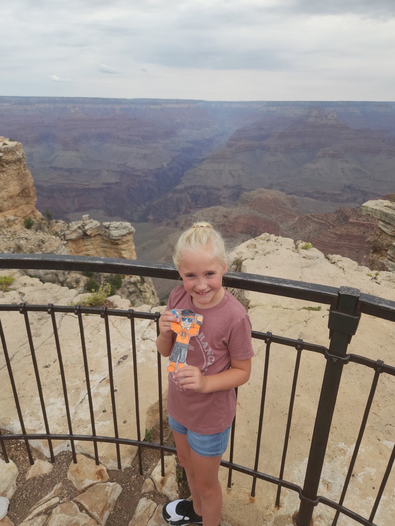 A young person stands on a cliff, holding a toy, with a metal railing. Below is a vast, mountainous landscape.