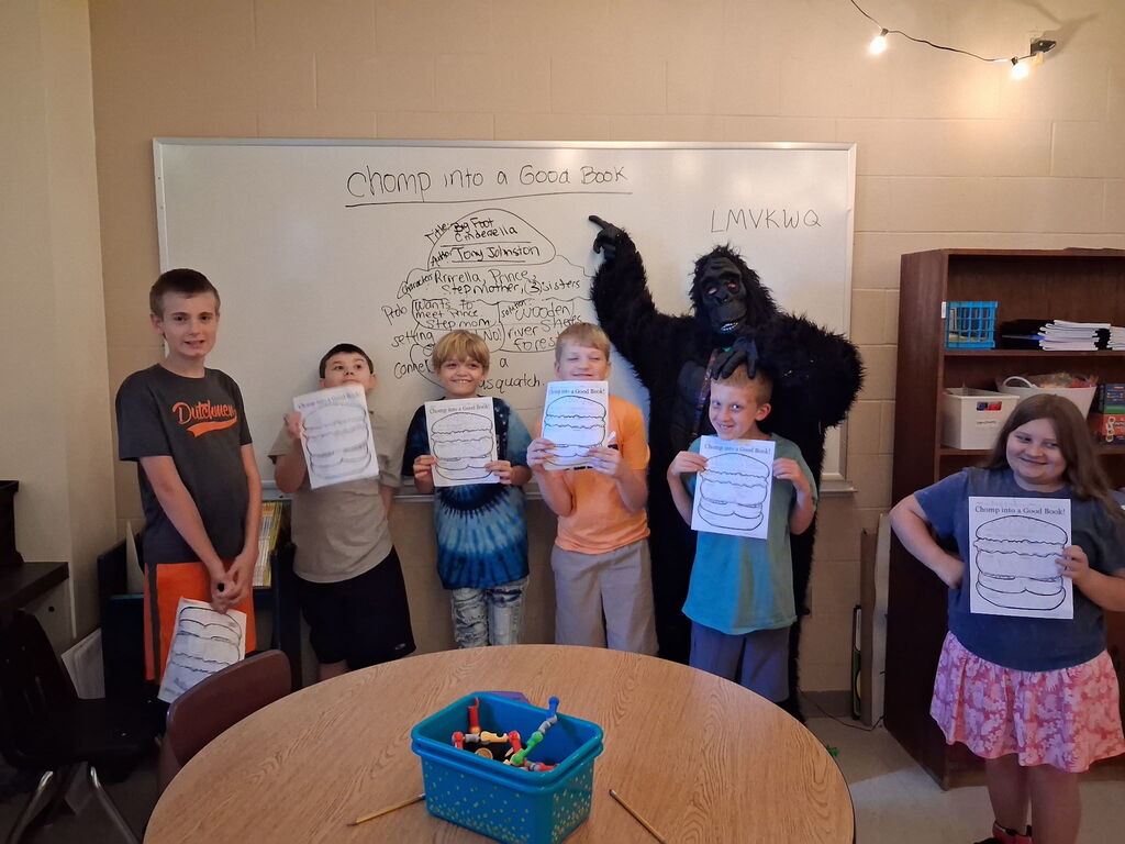 Children hold certificates; one is a gorilla mascot. They stand in a classroom, with a whiteboard, shelves, and a table.