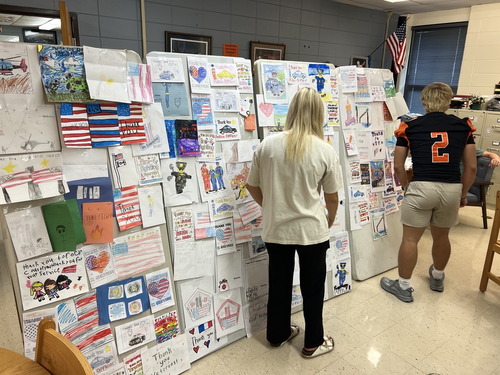Two people view a wall with many colorful drawings and cards attached, with one person wearing a number 2 jersey.