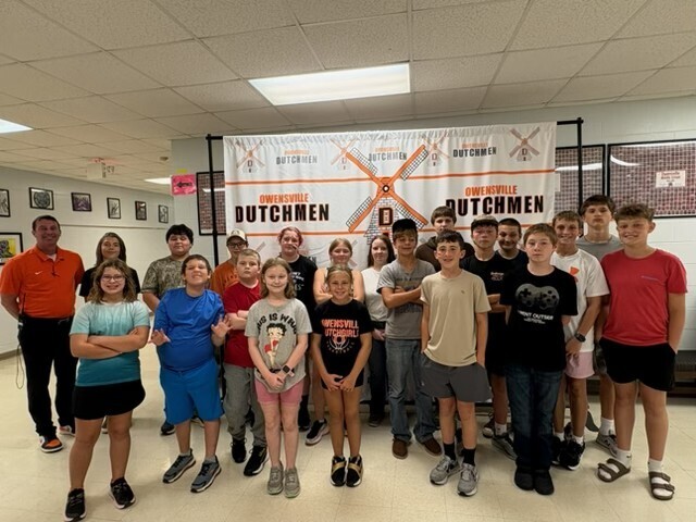 Group of children and adults pose for photo in classroom with "Dutchmen" banner in background.