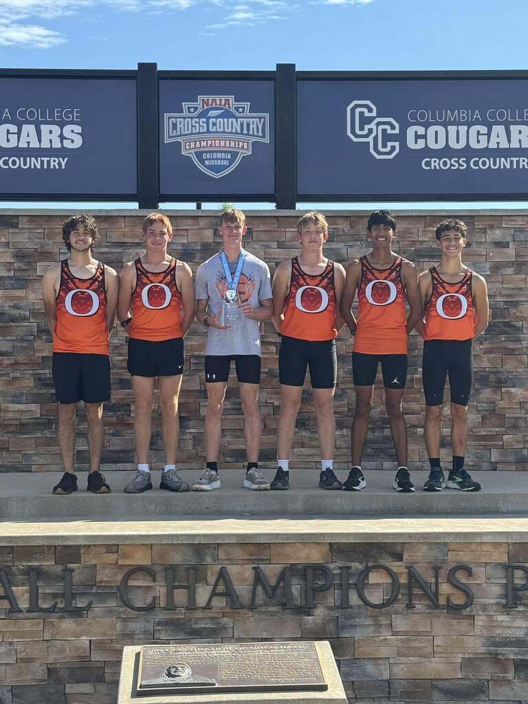A group of athletes stand on a podium. Five wear orange shirts with the letter "O" and black shorts.