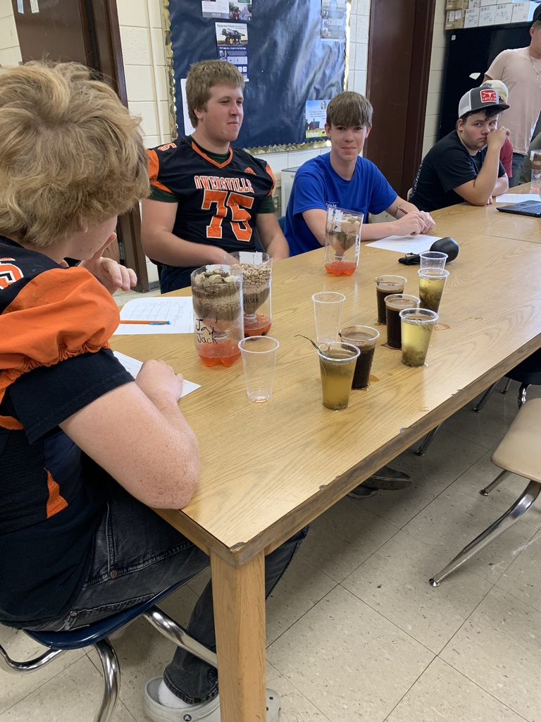 Five students wearing sports jerseys are seated around a table with drinks. A teacher sits nearby, looking at a student.