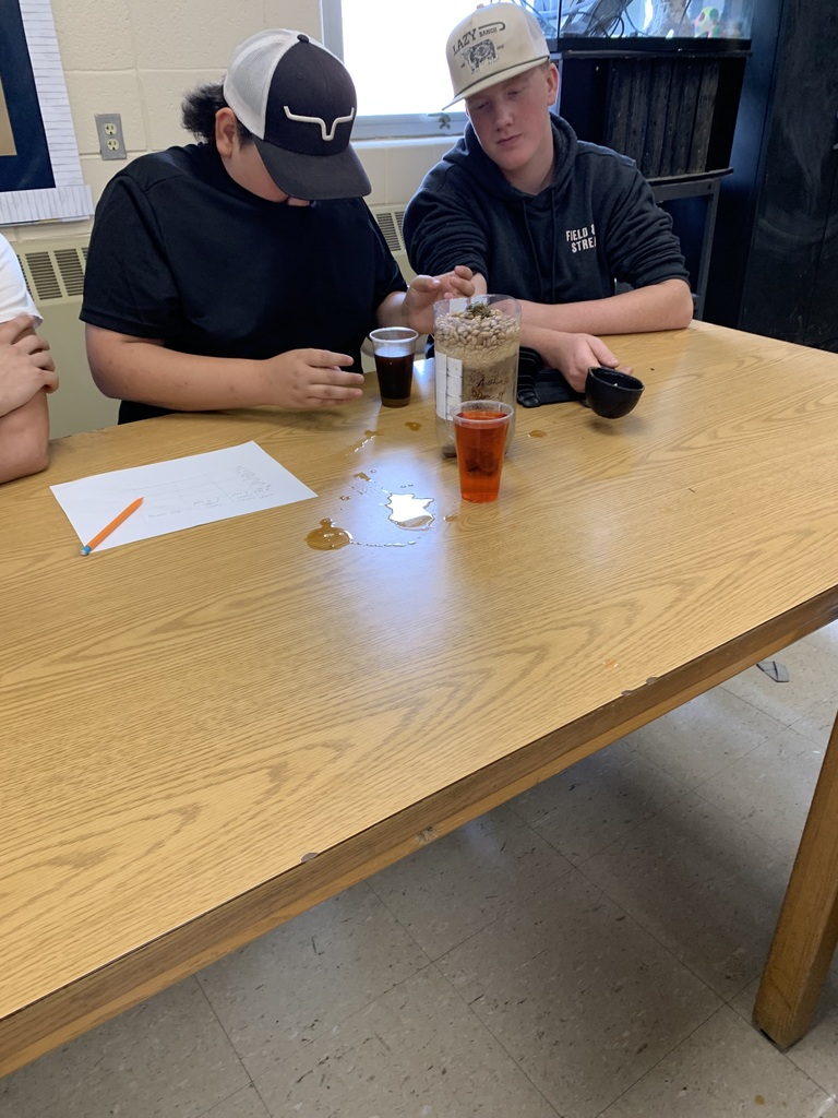 Two individuals wearing caps sit at a table, with one person handling a bottle and cups.