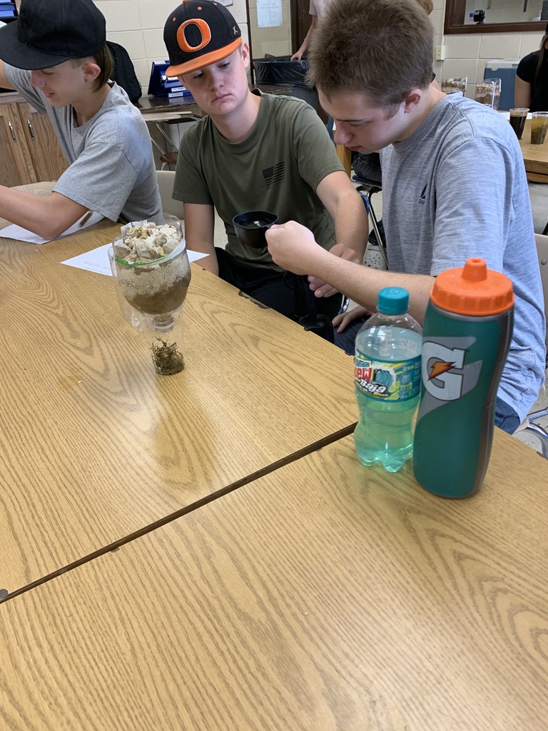 Three individuals seated at a table, one of them observing a glass container filled with a layered substance.