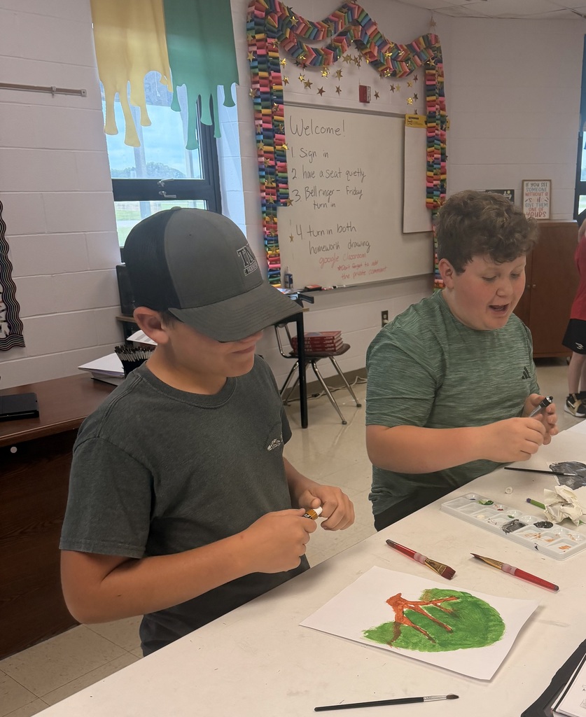 Two boys in a classroom, one with a hat, both engaged in art, brushes and paint visible.