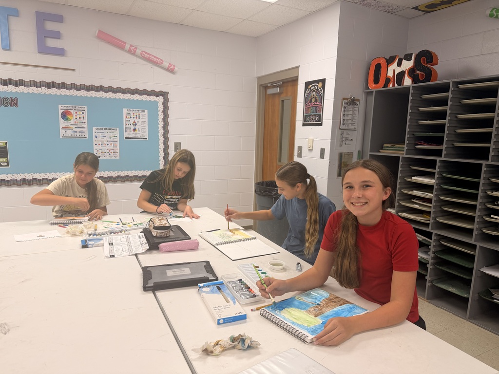 Four students in a classroom sit at a long table, working on art projects with various supplies.