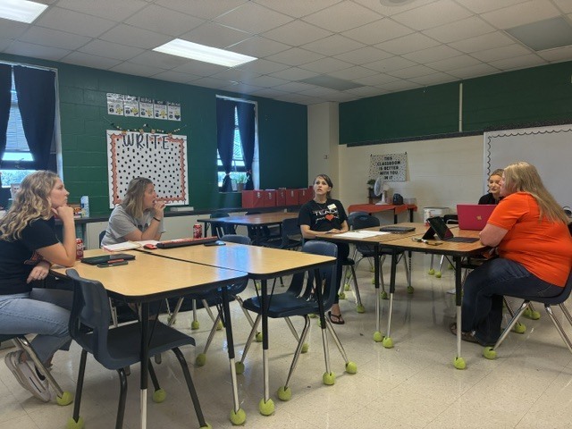 Five individuals seated around desks in a room with green walls, engaged in discussion. A whiteboard is present.