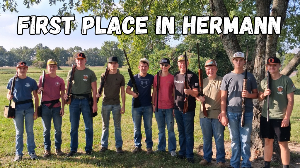 Group of individuals standing in a grassy field holding rifles. A large tree behind them. Text reads "FIRST PLACE IN HERMANN".