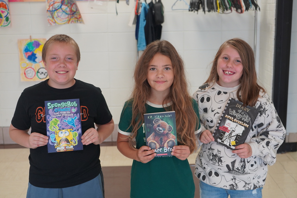 Three children pose for a photo. They hold books. A white wall is in the background.