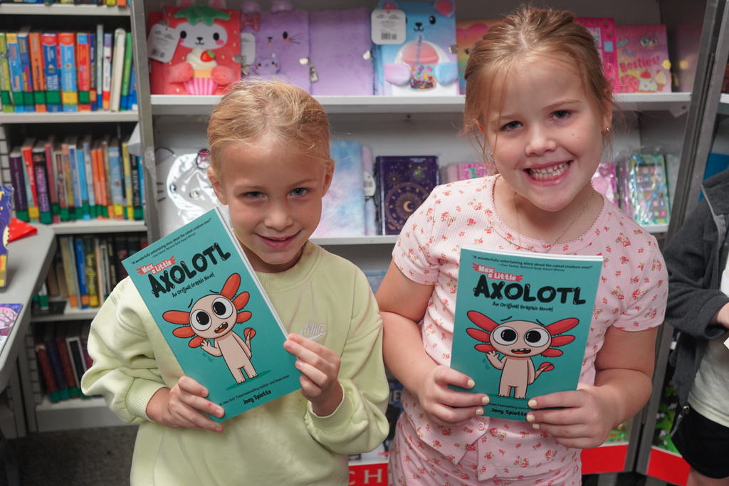 Two young girls hold books with colorful covers in a bright, colorful bookstore with shelves full of books.