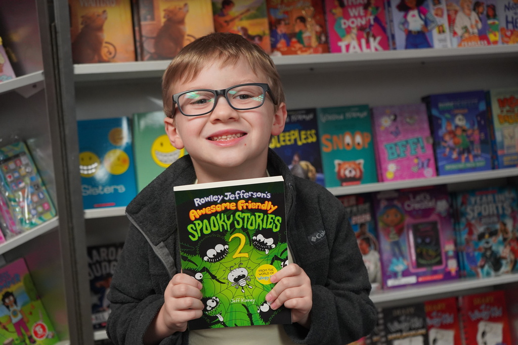 Child in glasses holds a book in a book store with many colorful books on shelves.
