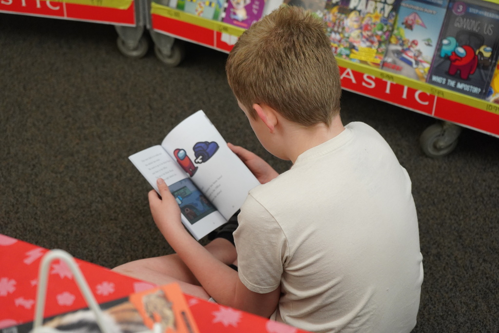 A boy reads a book with a colorful illustration while sitting on the floor in a bookstore.