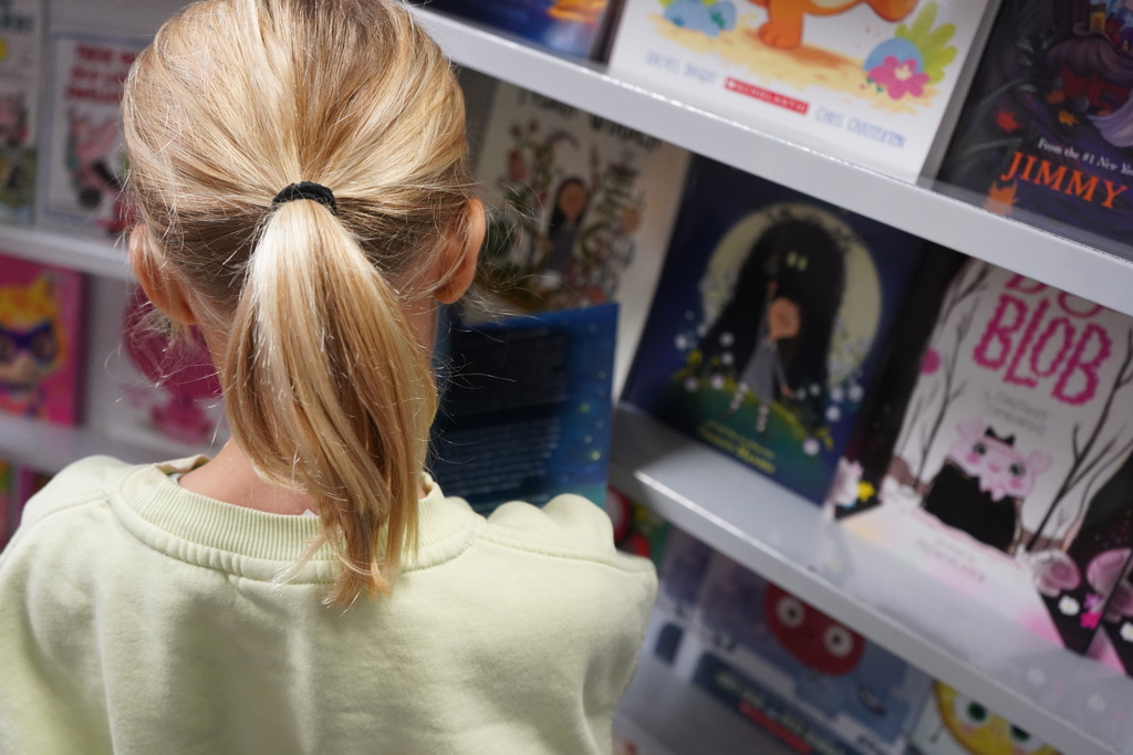 Child with a ponytail viewing books on a shelf. The books include titles like "Blob" and "Jimmy" among others.