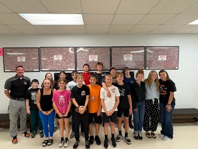 A group of children and adults pose for a photo in a classroom with white walls and framed pictures.