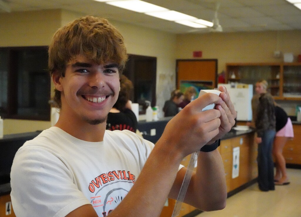 A man in a white shirt smiles and holds a gaming controller in a lab. Behind him, blurred figures are in a lab setting.