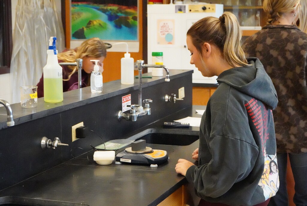 A woman in a hoodie stands at a countertop, looking at a scale, with two other people nearby.