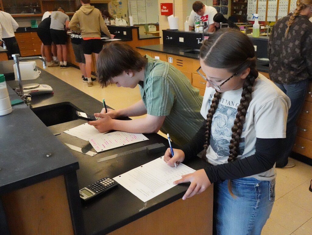 Students writing in a science lab. One student uses a calculator and the other writes with a pen.