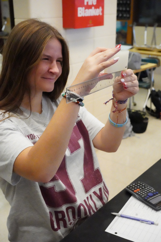 A girl holds a clear plastic ruler in a classroom. She wears a gray shirt with maroon letters and a number 42.