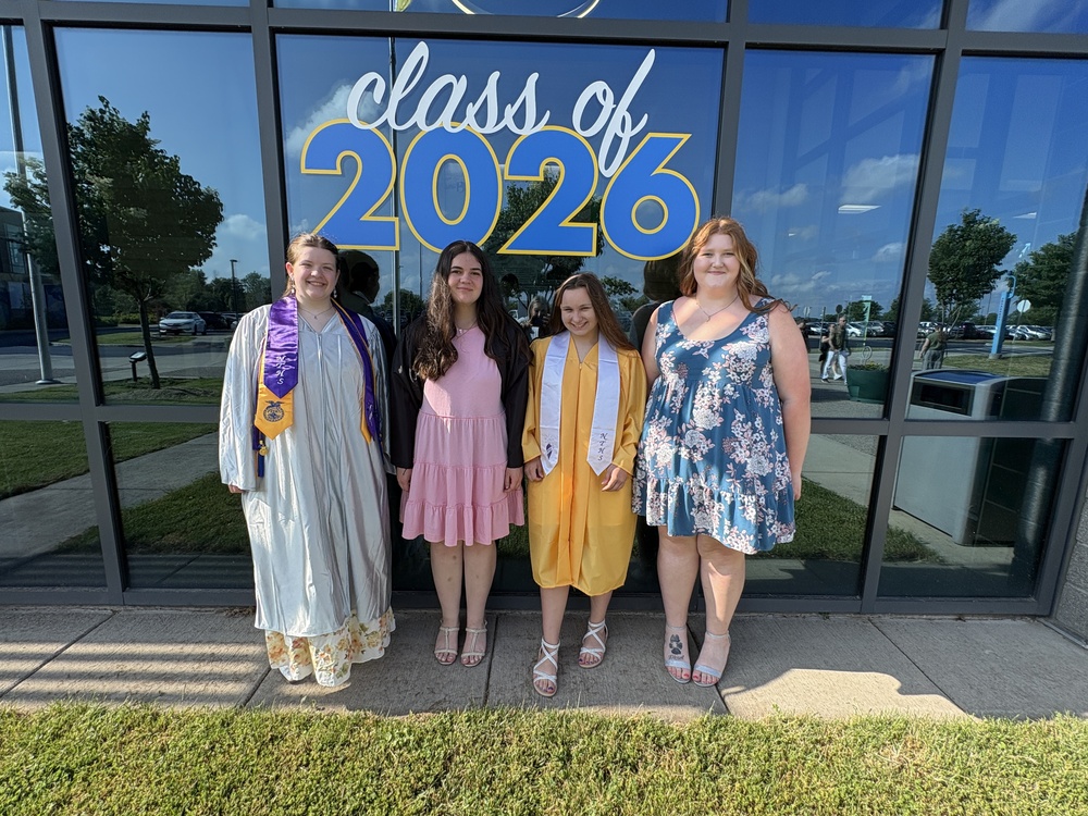 Four students stand in front of the class of 2026 sign with two in graduation gowns.