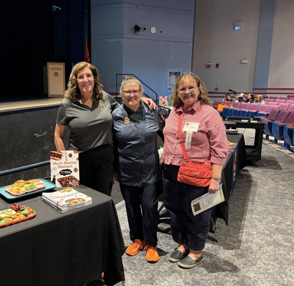 Three women stand together beside a food service display table at an educational event. The table features sample meals, promotional materials and signs highlighting school meal options, including National Chicken Sandwich Day and “March Meatball Madness.” The group is smiling for the photo in front of an auditorium stage, with additional event displays and attendees visible in the background.