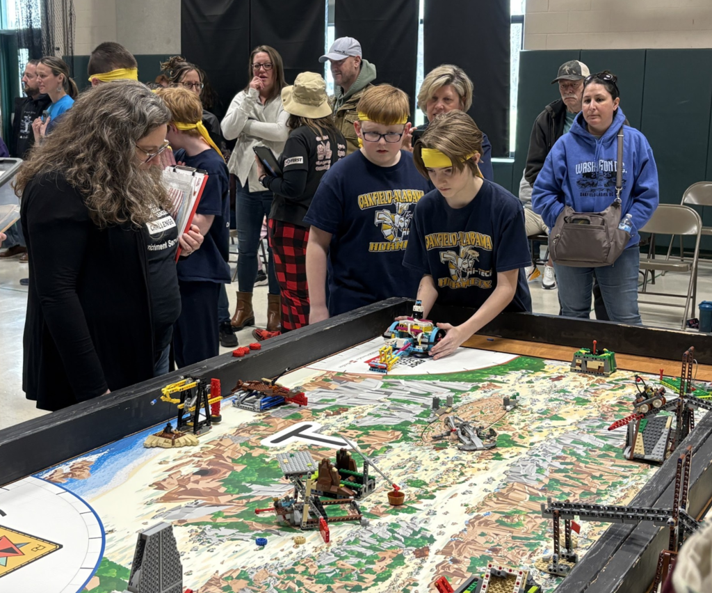 Two students during a robotic match setting up their robot to complete tasks while judges and parents watch in excitement.  