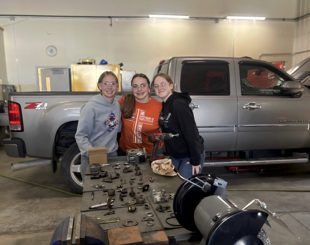 Three female students standing in the Diesel Mechanic Shop posing in front of a truck and tools.  