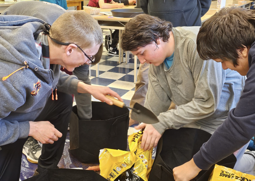 One student is holding a gardening tool, one is emptying soil into a pot and one is helping the student with the soil. 