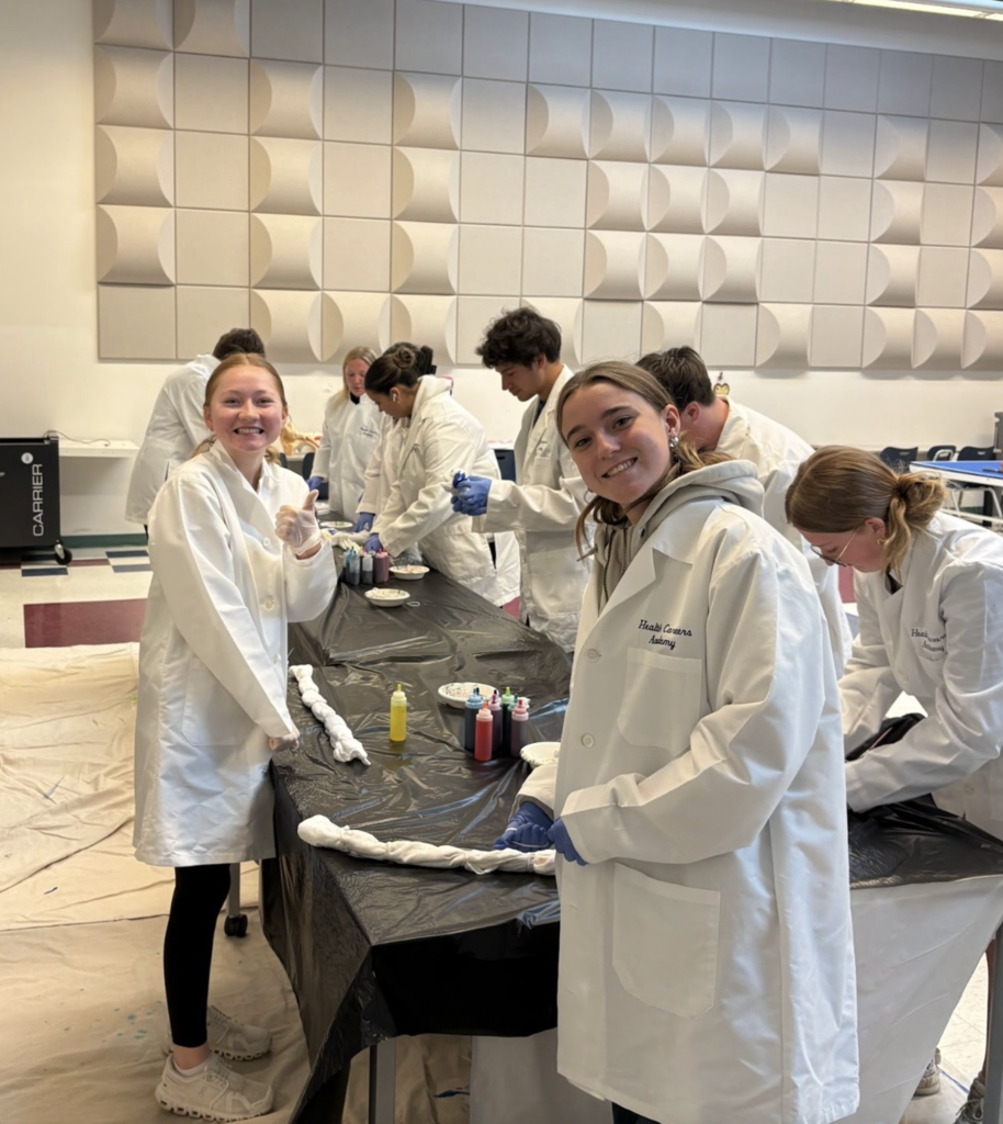 Students in Health Careers Academy lab coats work together on a tie-dye activity, twisting and dyeing fabric at tables covered in protective plastic. Wearing gloves and using colorful dye bottles, the students appear to be learning and collaborating in a hands-on classroom activity while creating custom tie-dye designs.