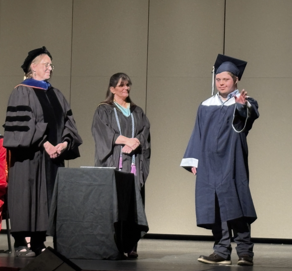 This photo captures a graduation ceremony moment on stage. A graduate wearing a cap and gown stands beside two faculty or staff members dressed in academic regalia.