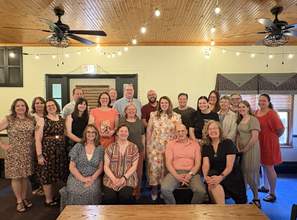 This photo shows a group of librarians gathered with an author for a commemorative picture at an annual dinner. Everyone is posed together indoors in a warmly lit venue with string lights and a rustic wooden ceiling, creating a welcoming atmosphere.