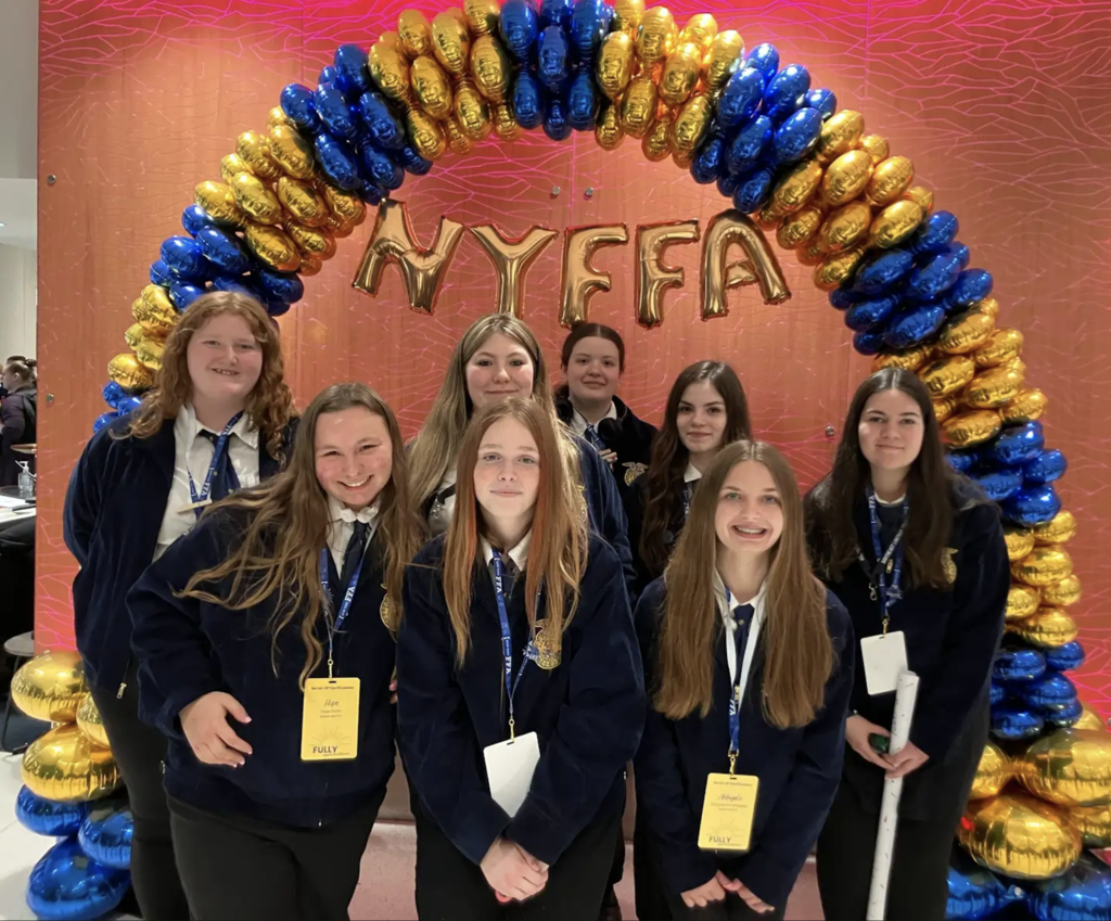 A group of students wearing official blue FFA jackets pose together in front of a large blue-and-gold balloon arch with gold letter balloons spelling “NYFFA.” The students are attending an FFA conference and are smiling for a group photo while wearing conference badges and lanyards. One student is holding a rolled poster or banner, adding to the event atmosphere.