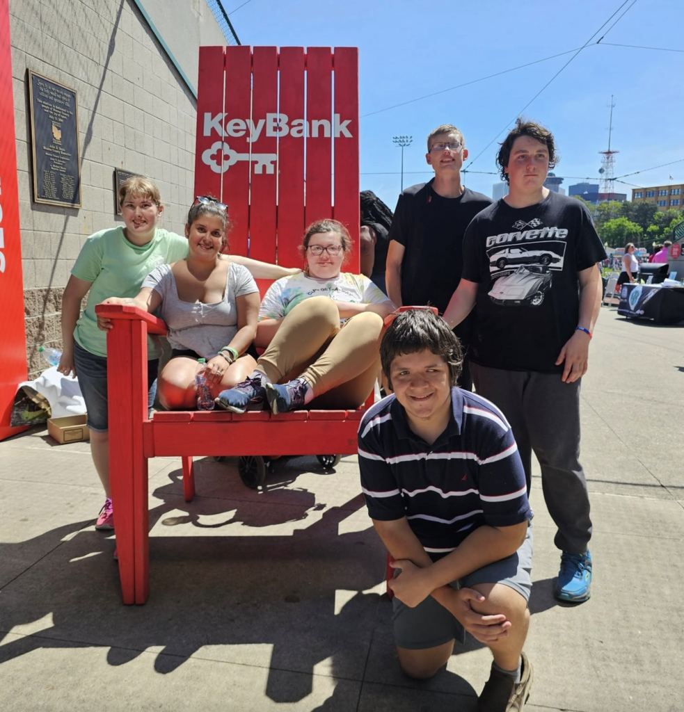 Two students are sitting in the oversized chair, while others stand beside it and one student kneels in front.