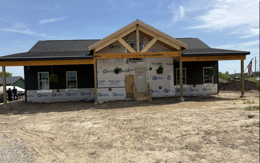 A nearly completed house sits on a dirt lot with its roof, windows, and front porch already installed. Construction wrap still covers parts of the exterior, showing that work is still underway.