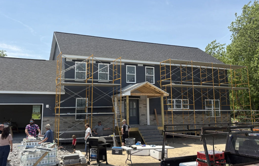 A large two-story house is under construction, with scaffolding surrounding the front of the building as work continues on the exterior. Workers and visitors gather outside near construction materials, tools, and equipment in front of the nearly completed home.