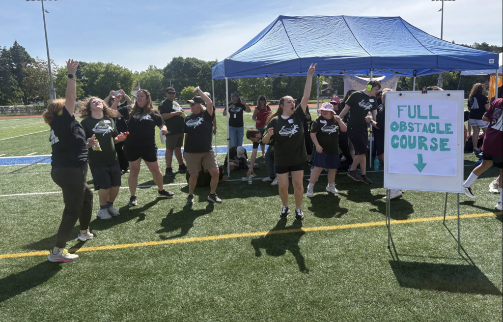 Students and staff take a break from activities to enjoy an impromptu dance party during lunch. Gathered near a tent on the athletic field, they danced, laughed, and celebrated together while enjoying the sunny day.