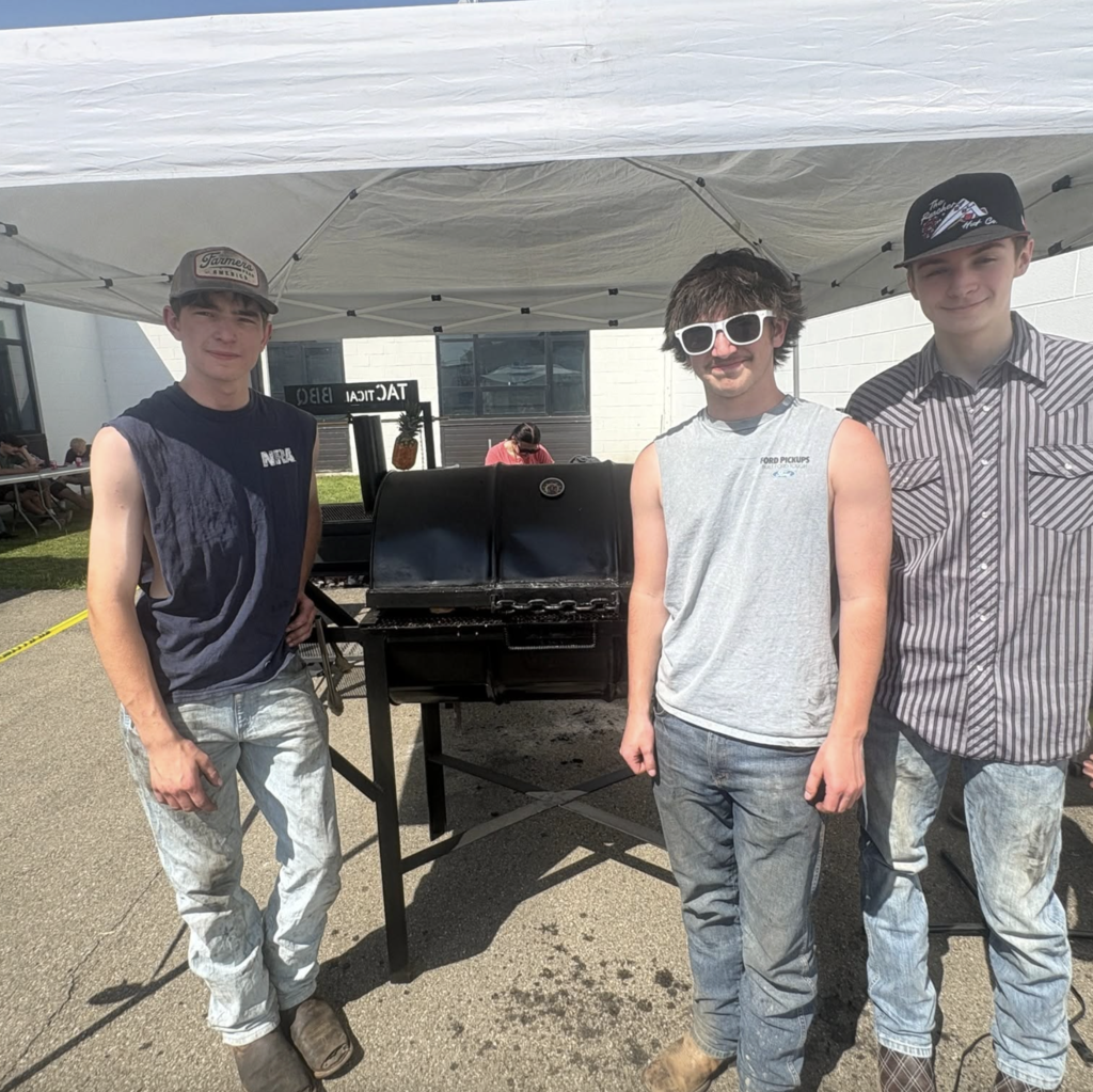 Three students stand beside a grill under a canopy tent during an outdoor event. The group poses for a photo while helping prepare food, showcasing their teamwork and participation in the activity.