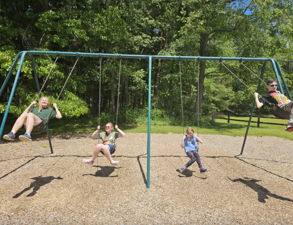 Four children enjoy a sunny day on the playground as they swing on a swing set surrounded by trees. Smiles and laughter fill the scene as they take turns soaring through the air and spending time together outdoors.