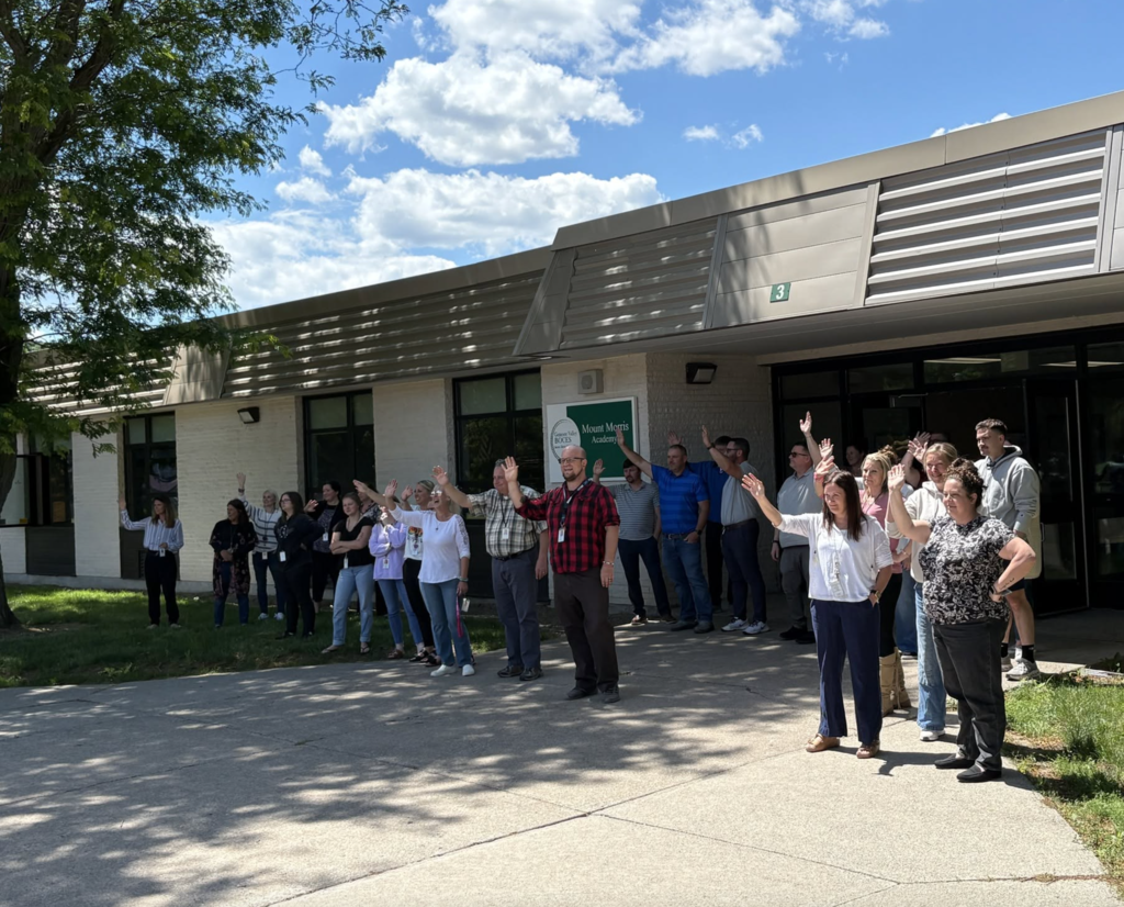 Teachers and staff standing outside the building waving good bye. 