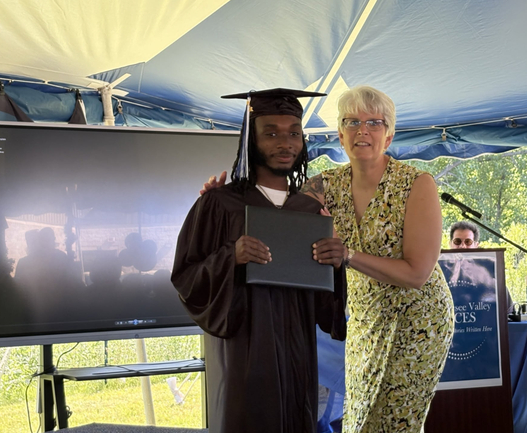 A student stands on stage with a certificate with a teacher standing side by side happy.  
